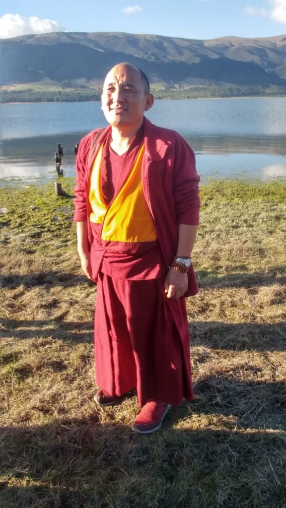 Monje budista de pie junto a un lago, con montañas de fondo.